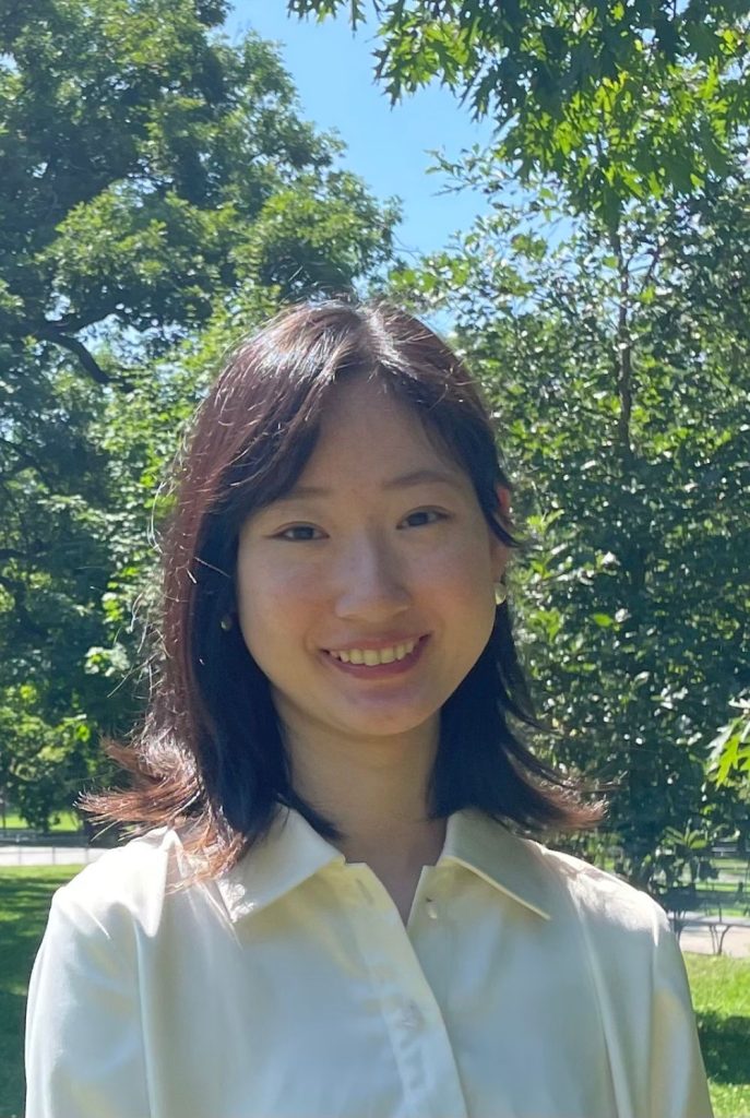 A person with shoulder-length dark hair is smiling while standing outdoors in a sunlit area with trees in the background.