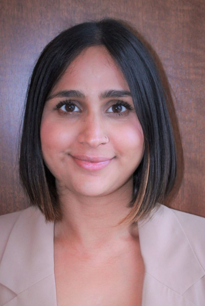 A person with shoulder-length dark hair, wearing a beige blazer, is standing in front of a wooden background.