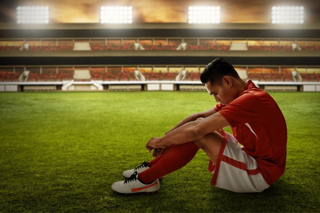 A soccer player sits alone on the field in an empty stadium, head lowered — the aftermath of a difficult game, when performance anxiety often has its strongest impact