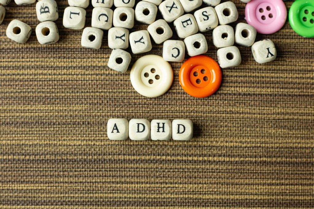 Wooden letter blocks spell “ADHD” below scattered blocks and colorful buttons on a brown striped fabric background, visually representing the question: Is ADHD Assessment Covered by OHIP?